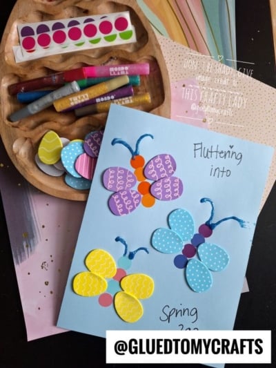A blue card with colorful paper butterfly crafts, labeled “Fluttering into Spring 2023,” sits beside decorative stickers, paint markers, and egg-shaped cutouts. A tray with craft supplies is visible, plus the @GLUEDTOMYCRAFTS watermark.