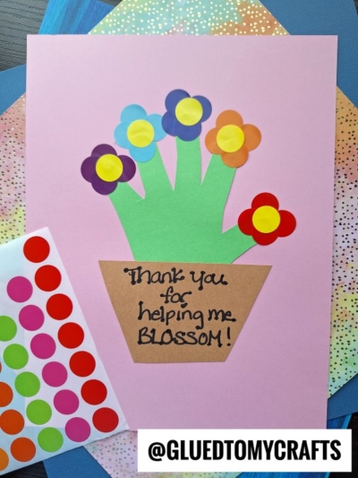 A handmade card with paper cutouts forming a flower pot and flowers, reading Thank you for helping me blossom! Coding label crafts, colorful sticker dots, and craft papers surround the card. @GLUEDTOMYCRAFTS is printed at the bottom.