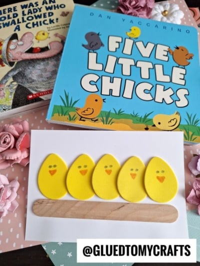 A craft project features five yellow chick shapes glued to a wooden stick, displayed on white paper. Behind it are two children’s books with chick illustrations, surrounded by pink and white decorative flowers.