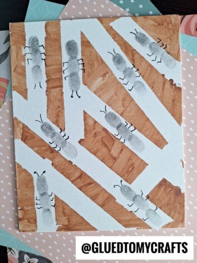 A canvas with brown and white geometric shapes and black fingerprint ants, with six legs and antennae drawn in black, on top. The image has a watermark that says @GLUEDTOMYCRAFTS.