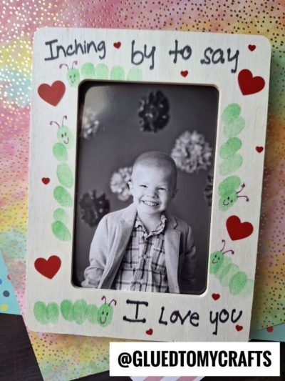 A decorated wooden picture frame with green caterpillars and red hearts surrounds a black-and-white photo of a smiling child. The frame reads, Inching by to say I love you. The handle @GLUETOMYCRAFTS is at the bottom.