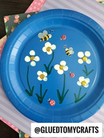 A blue paper plate decorated with painted white flowers, bees, and ladybugs, placed on patterned paper sheets. The flowers have yellow centers, green stems, and leaves. The bees and ladybugs are drawn with simple details.