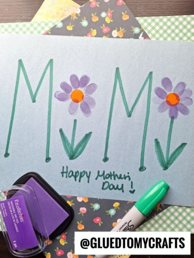 A handmade Mother’s Day card spells “MOM” with green letters, using purple flowers with orange centers as the “O.” There’s a green marker, purple ink pad, patterned papers, and text: “Happy Mother’s Day!” and “@GLUEDTOMYCRAFTS.”.