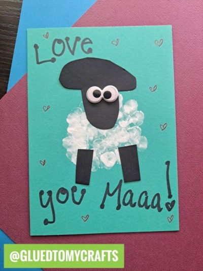 A handmade card features a sheep crafted from black paper shapes and white paint for its wool, with googly eyes. The text reads Love you Maaa! and small hearts decorate the teal card. @GLUEDTOMYCRAFTS is at the bottom.