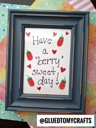 A framed sign with hand-drawn strawberries, red hearts, and the message Have a berry sweet day! on white paper. The Instagram handle @GLUEDTOMYCRAFTS is displayed at the bottom. Colorful papers are in the background.