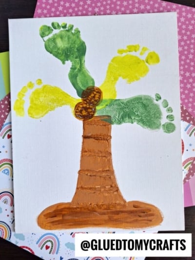 A painting of a palm tree made with green and yellow painted footprints as leaves and two brown painted coconuts, with a brown trunk on a white canvas. The handle @GLUEDTOMYCRAFTS is at the bottom right.