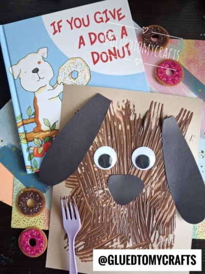 A children’s book titled If You Give a Dog a Donut with a charming fork painted dog craft made of brown paint, googly eyes, and black paper ears and nose, plus toy donuts and a purple fork on a colorful background. @GLUEDTOMYCRAFTS.