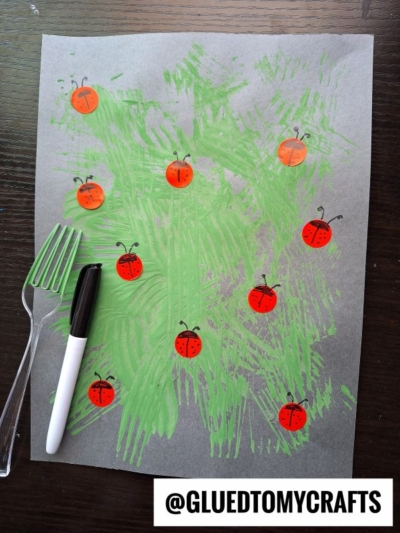 A grey paper with green paint streaks, made using a plastic fork, is decorated with red and orange ladybugs drawn with a black marker. A coding label crafts touch is added, with a black marker and plastic fork shown beside the paper. @GLUEDTOMYCRAFTS appears at the bottom.