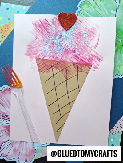 A handmade ice cream cone craft with a brown paper cone, pink painted ice cream, blue glitter, and a red glitter heart on top. An orange fork is beside it, and colorful flower cutouts decorate the background.