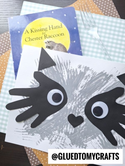 Children’s book “A Kissing Hand for Chester Raccoon”—perfect for back to school story time—pairs with DIY raccoon art made from black paper, googly eyes, and gray paint, set on patterned paper with a plastic fork. @GLUEDTOMYCRAFTS watermark at bottom.