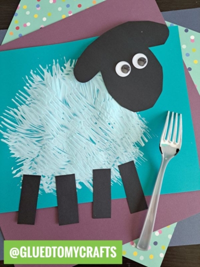 A sheep craft made from a blue painted body, black paper head and legs, googly eyes, and a plastic fork on a green background. Decorative polka dot papers are layered underneath.