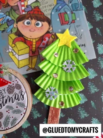 A handmade Christmas tree ornament made from green paper, a yellow felt star, sequins, and snowflake decorations on a popsicle stick, with a cartoon girl, cupcake liner card crafts, and Christmas cards in the background.