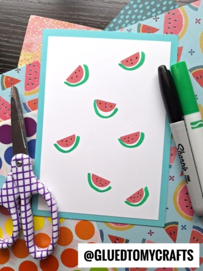 A handmade card decorated with small watermelon slice illustrations lies on colorful patterned paper, perfect for coding label crafts. Nearby are a pair of purple scissors and two Sharpie markers. The handle @GLUEDTOMYCRAFTS appears at the bottom.