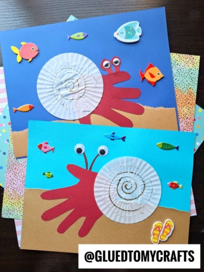 Two handmade crab crafts using red cut-out handprints and white cupcake liners for shells, decorated with googly eyes and surrounded by colorful fish stickers on blue backgrounds, with @GLUEDTOMYCRAFTS in the corner.