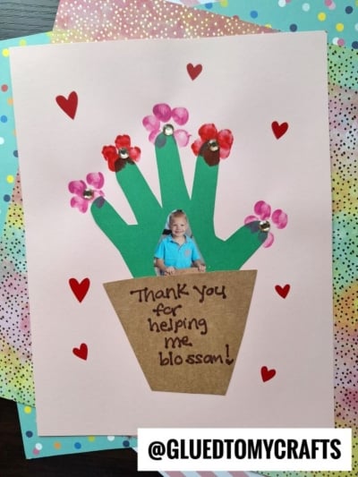 A handmade card features a flower pot with green paper stems, red and pink flower prints topped with sequins, a child’s photo as the flower center, red heart stickers, and the message “Thank you for helping me blossom!”.