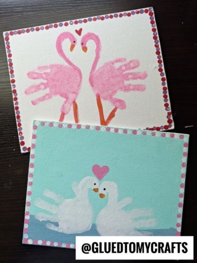 Two paintings made with handprints: the top one shows two pink flamingos forming a heart shape with their necks, and the bottom one shows two white swans with a pink heart above them. Both have dotted borders.
