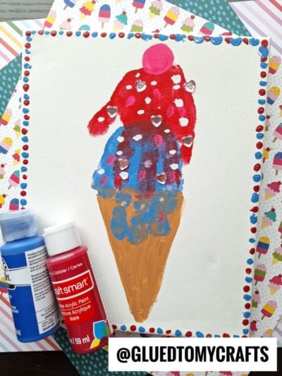 A painting of an ice cream cone with two scoops—pink on top with white dots and blue with red and white dots—on a white canvas, surrounded by colorful patterned paper and two paint bottles. @GLUEDTOMYCRAFTS is in the corner.