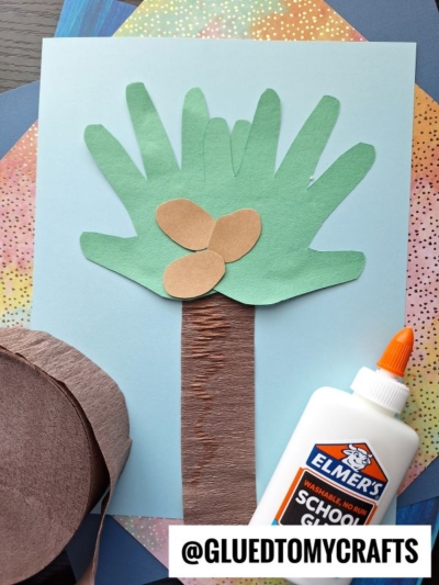A paper craft coconut tree made with green, brown, and light blue paper, with a roll of brown crepe paper and a bottle of Elmer’s School Glue beside it. The username @GLUEDTOMYCRAFTS is at the bottom.