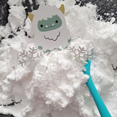 A cute paper yeti with snowflakes and a blue spoon sits on a pile of white, fluffy faux snow. The Instagram handle @GLUEDTOMYCRAFTS appears in bold text at the bottom right.