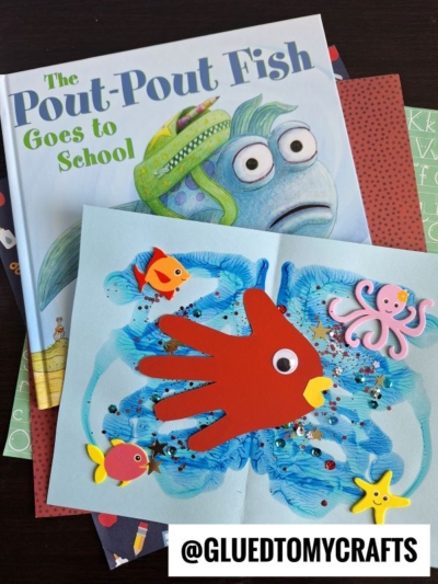 A children’s book titled The Pout-Pout Fish Goes to School sits beneath an ocean-themed craft with a red handprint fish, foam sea creatures, glitter, and blue paint—perfect for back to school story time. The handle @GLUEDTOMYCRAFTS appears at the bottom.