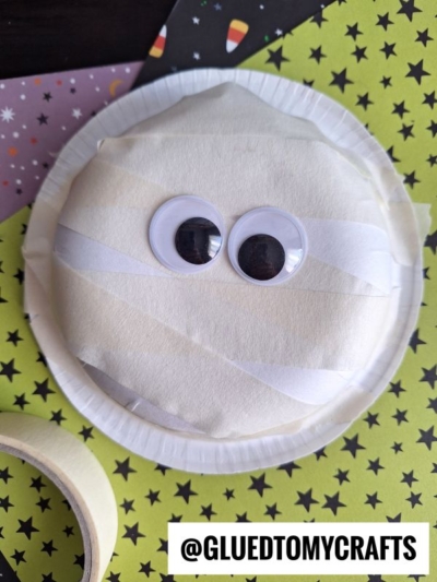 A paper plate mummy craft with large googly eyes, wrapped in white paper strips, sits on a green star-patterned sheet with washi tape nearby—perfect inspiration for fans of paper bowl crafts. The text @GLUEDTOMYCRAFTS appears at the bottom.