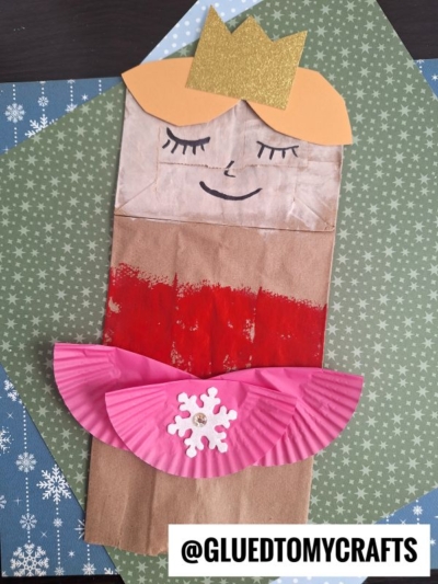 A paper bag puppet of a ballerina with a gold crown, orange hair, red dress, and pink cupcake liner tutu, decorated with a white snowflake. The puppet is on green and blue snowflake-patterned paper. Text: @GLUEDTOMYCRAFTS.