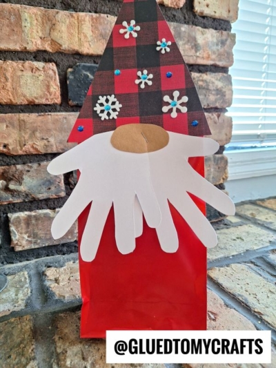 A handmade gnome craft with a plaid hat, paper snowflakes, a round nose, and white handprint-shaped beard, placed on a red paper bag against a brick wall. The text @GLUEDTOMYCRAFTS appears at the bottom.