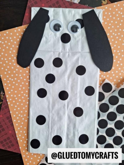 A white paper bag decorated as a Dalmatian dog with black paper ears, black spots, large googly eyes, and lying on patterned orange and red paper—perfect for coding label crafts. The handle @GLUEDTOMYCRAFTS appears at the bottom.