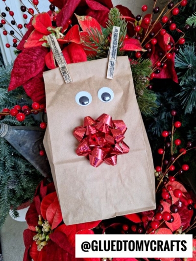 A brown paper bag decorated as a reindeer with googly eyes, a red gift bow for a nose, and two clothespins as antlers, set among red and green holiday foliage. The text @GLUEDTOMYCRAFTS is at the bottom.