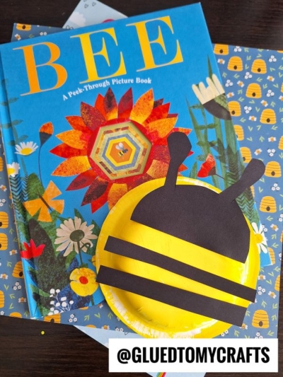 A children’s book titled “BEE” lies on a floral-patterned surface with a yellow and black paper plate bee craft on top. The bee has black stripes and antennae. @GLUEDTOMYCRAFTS is labeled in the corner.