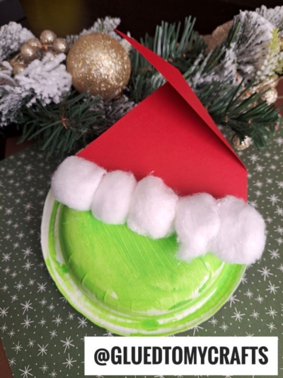 A green paper plate decorated as the Grinch’s face with cotton balls and a red paper Santa hat, set against a festive background with pine branches and gold ornaments—a fun twist on classic paper bowl crafts. Text reads @GLUEDTOMYCRAFTS.