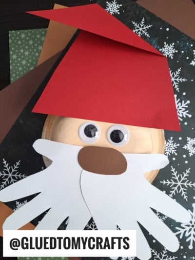 A Santa face craft made with a red paper hat, large googly eyes, and a white paper beard shaped like two handprints—perfect for fans of paper bowl crafts—displayed on snowflake and brown paper backgrounds.