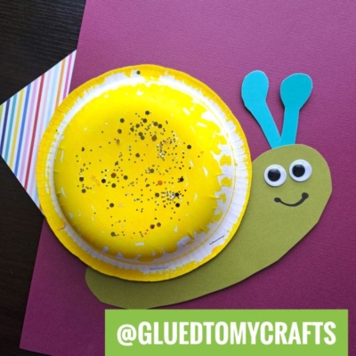 A snail craft made from a yellow-painted paper plate for the shell, with a green paper body, two blue antennae, googly eyes, and a drawn smile, placed on a pink background. Text reads @GLUEDTOMYCRAFTS.