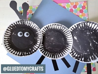 Three black painted paper plates are arranged to resemble a sheep, with googly eyes on one plate and cut-out black paper legs and antennae on colorful scrapbook paper. @GLUEDTOMYCRAFTS is written at the bottom left.