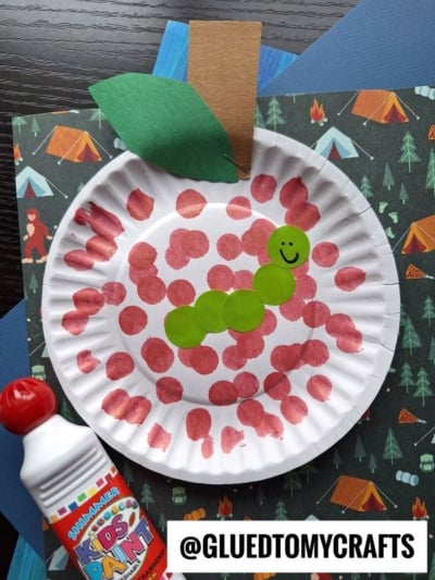 A paper plate painted with red dots to resemble an apple, featuring a green paper leaf, a brown stem, and a smiling green caterpillar made from painted circles—perfect for a fun Hungry Caterpillar craft. Bottled kids paint and blue paper are nearby.