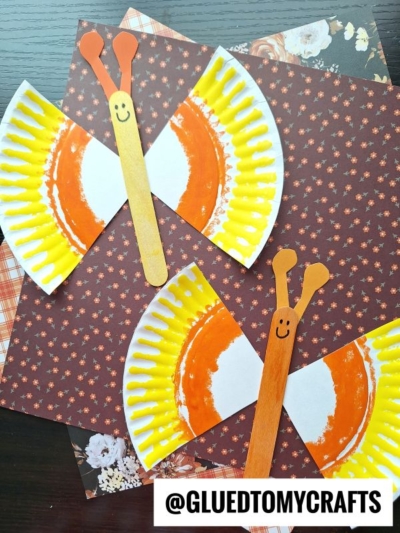 Two butterfly crafts made from paper plates painted orange and yellow, with wooden sticks as bodies and paper antennae, are displayed on floral-patterned paper. A @GLUEDTOMYCRAFTS watermark is at the bottom.