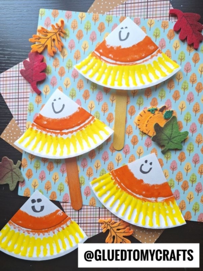 Four paper plate crafts shaped like smiling candy corn with painted orange and yellow edges, arranged on patterned paper with felt leaves. One has a craft stick handle. The image is labeled @GLUETOMYCRAFTS.