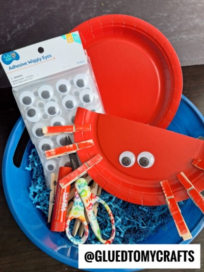 A blue basket holds red paper plates, adhesive googly eyes, clothespins, glue, scissors, and shredded blue paper, with a finished red crab craft featuring googly eyes and clothespin legs. @GLUEDTOMYCRAFTS label is visible.