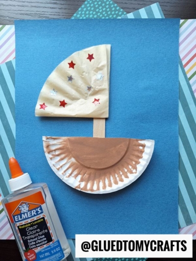 A kids sailboat craft made with a popsicle stick, a painted paper plate, and a decorated paper sail on blue paper. An Elmer’s glue bottle is on the left, and the handle @GLUEDTOMYCRAFTS is at the bottom.