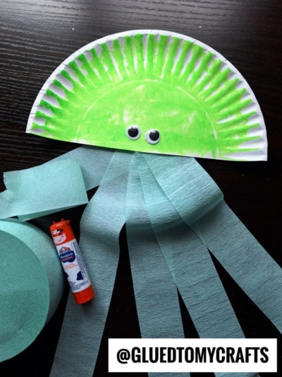 A green paper plate jellyfish craft with googly eyes and light blue crepe paper streamers for tentacles—perfect for those searching for fun crepe paper craft ideas. A glue stick, extra crepe paper, and @GLUEDTOMYCRAFTS are featured at the bottom.