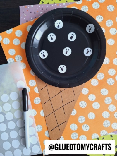 A black paper plate with white ghost faces is placed on brown paper cut into a cone shape to resemble ghost ice cream, surrounded by colorful patterned papers, a coding label, a black marker, and white sticker dots.