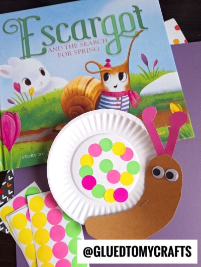 A children’s book titled Escargot and the Search for Spring is displayed behind a snail craft made with a paper plate, colored dot stickers, and construction paper on a purple surface—perfect inspiration for fun label crafts. @GLUEDTOMYCRAFTS is written below.