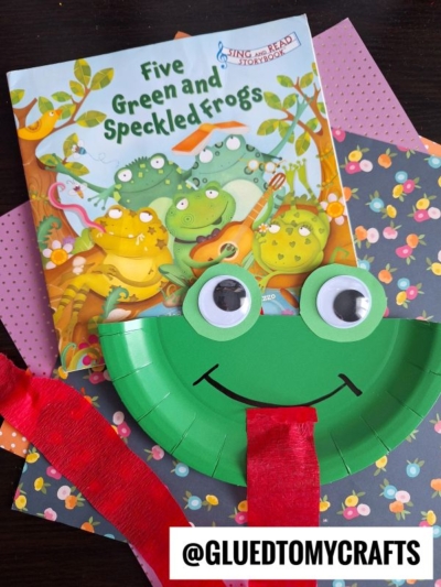 A children’s book titled Five Green and Speckled Frogs lies on colorful paper. In front is a frog craft made from a green paper plate, big googly eyes, and a red tongue—fun crepe paper craft ideas by @gluedtomycrafts are shown.