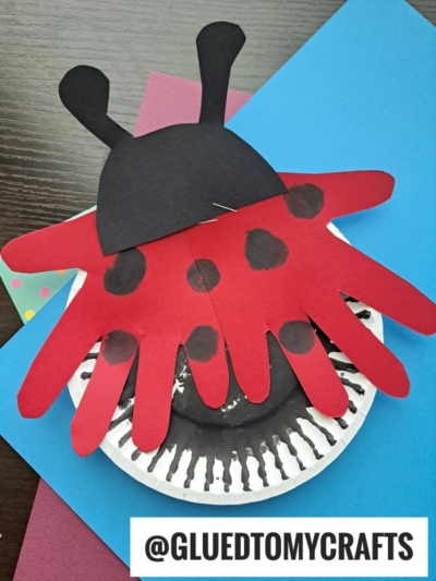 A ladybug craft made from red paper handprints with black spots for wings, a black paper head with antennae, and a white paper plate with black painted details, displayed on blue and pink paper.