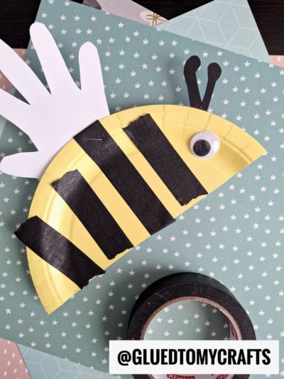 A bee craft made from a yellow paper plate with black stripes, a white paper handprint for wings, a googly eye, and black pipe cleaner antennae. Black washi tape and patterned paper are in the background.