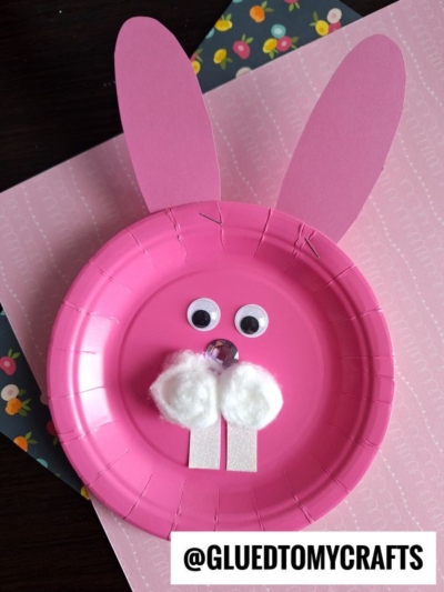 A pink paper plate decorated as a bunny with pink paper ears, googly eyes, a pom-pom nose, cotton ball cheeks, and white paper teeth. Background includes floral and pink patterned paper.