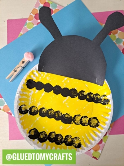 A paper plate painted yellow and black to resemble a bee with black paper antennae is displayed on colorful paper, next to a clothespin with a pink pom-pom. GLUEDTOMYCRAFTS is written in a green box at the bottom.