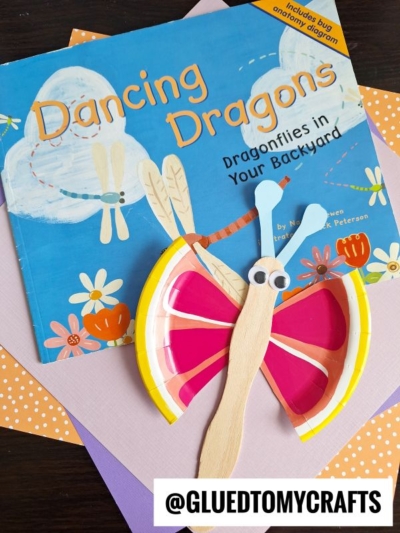 A childrens book titled Dancing Dragons: Dragonflies in Your Backyard is displayed with a dragonfly craft made from a paper plate and craft sticks. The background has pastel papers and @GLUEDTOMYCRAFTS text.