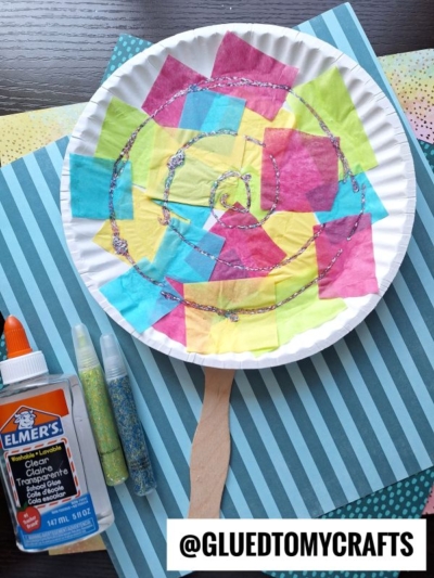 A paper plate decorated with colorful tissue paper squares and glitter glue, attached to a craft stick, sits on striped paper next to Elmer’s glue and two tubes of glitter. The handle @GLUEDTOMYCRAFTS is at the bottom.