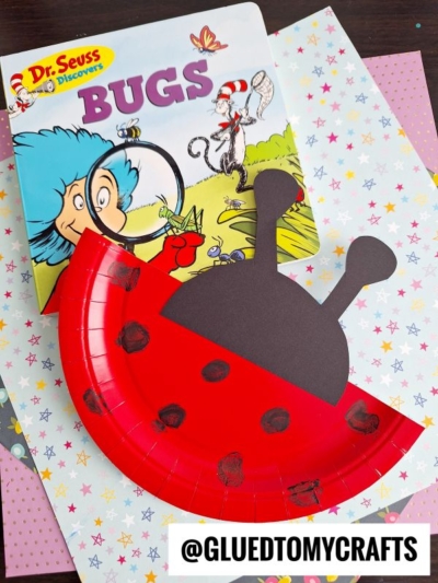 A Dr. Seuss Bugs book is next to a red paper plate ladybug craft with black spots and antennae, displayed on colorful star-patterned paper. The handle @GLUEDTOMYCRAFTS is visible at the bottom.